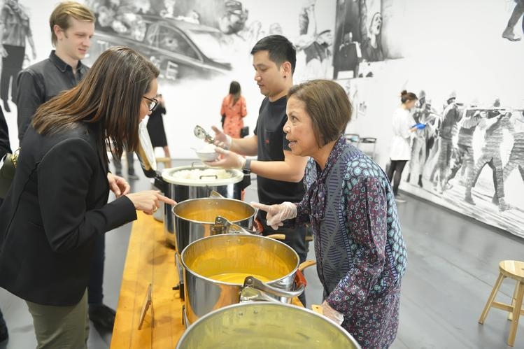People serving curry soup to guests while an artists draws on the wall in the background