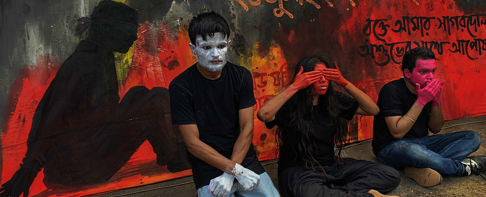Three people sit in front of a red and orange mural with text and the silhouette of a figure sitting, gagged and hands bound. The three people in the picture have painted their hands and faces in white, orange, and pink.