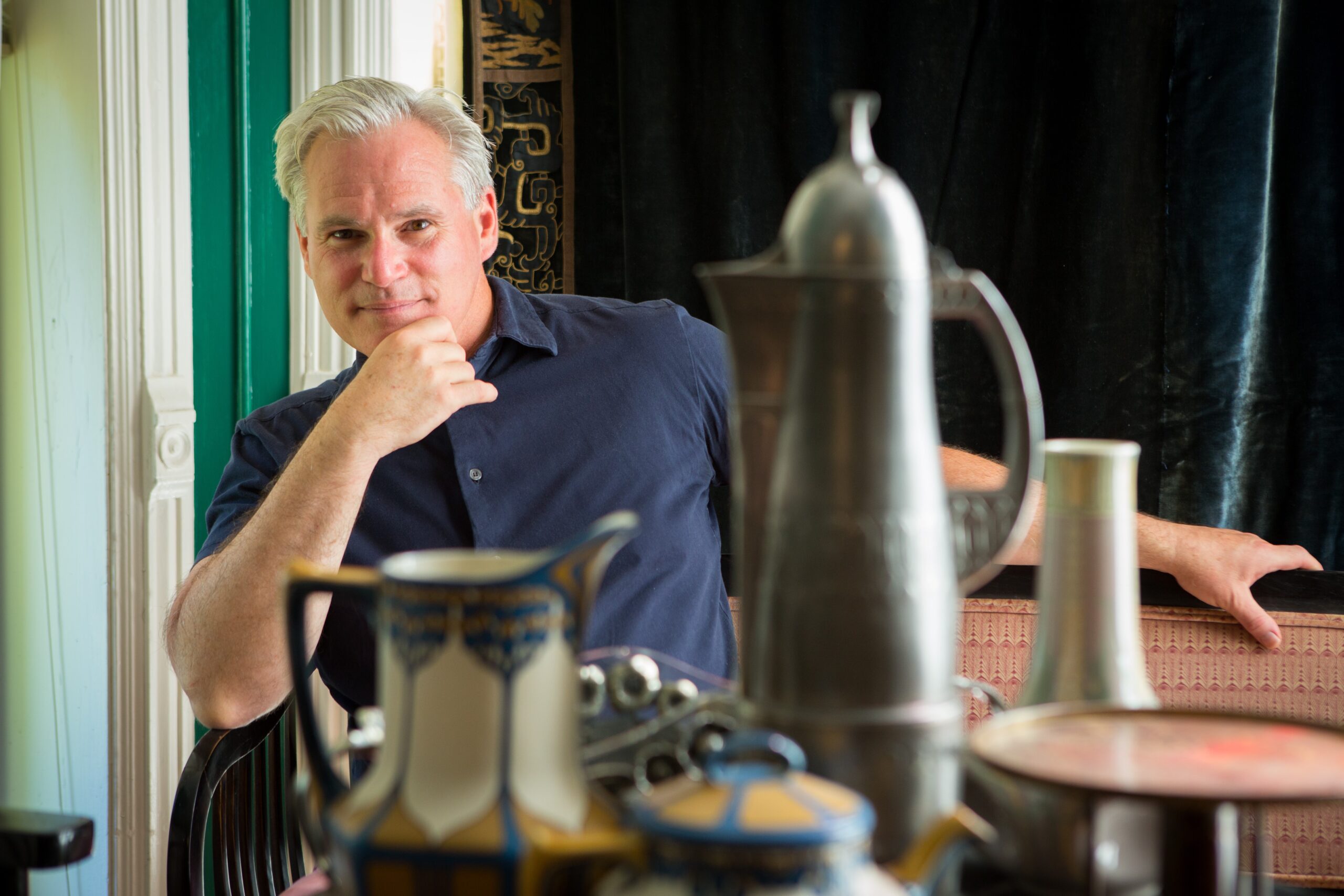 Photograph of Curator Jonathan D. Katz in a blue collared shirt. He is seated behind a silver tea pot and small carafe of cream.